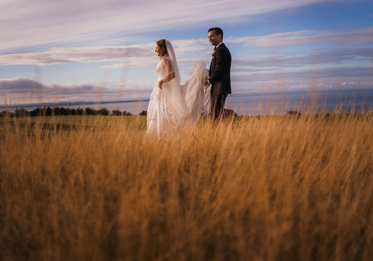 Groom holds brides train as they walk through grass together with sea in background